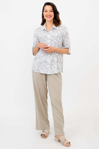 Full body view of a woman in a light patterned blouse and beige pants, smiling with hands together