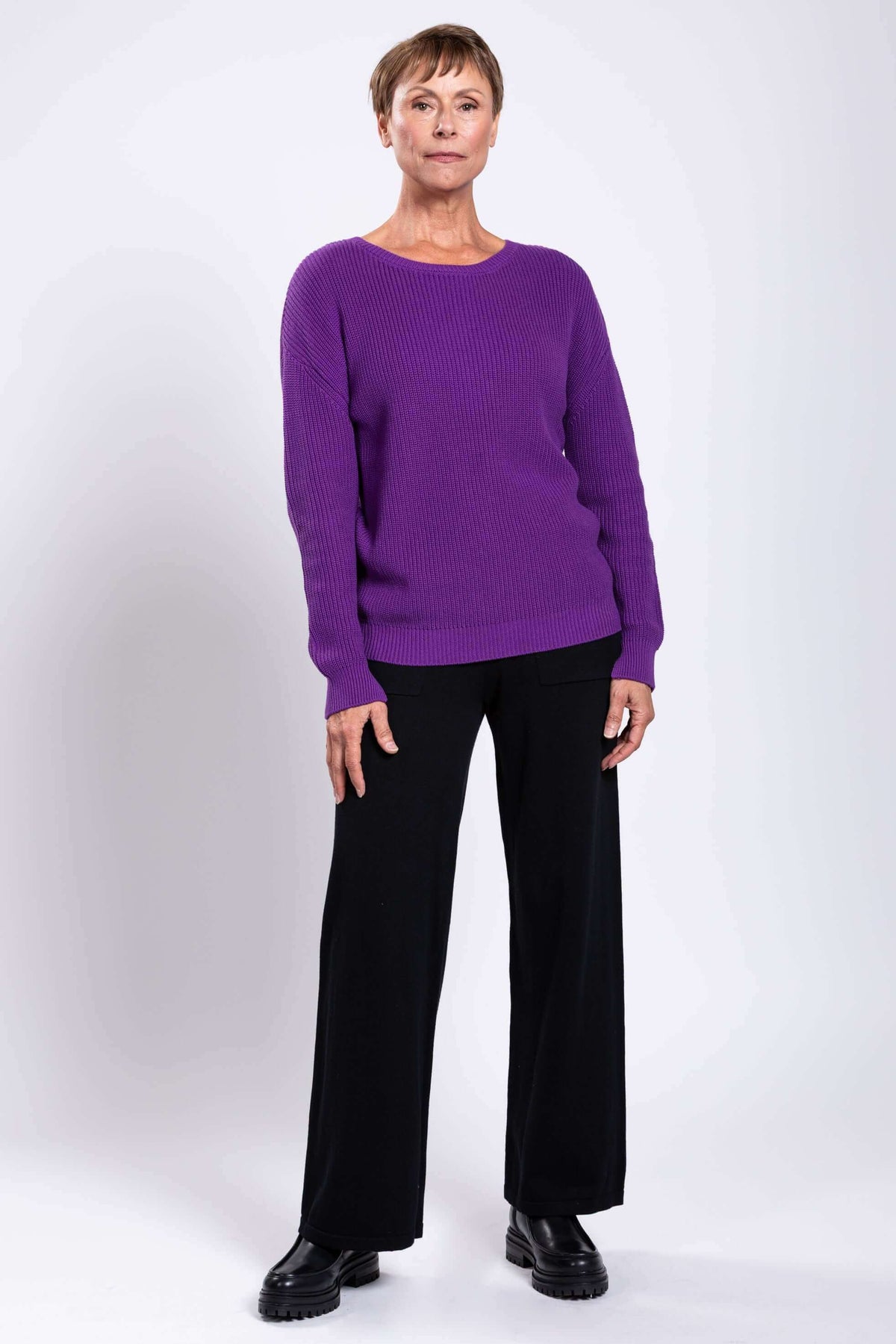 Woman in purple sweater and black pants, relaxed pose, light background