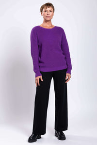Woman in purple sweater and black pants, relaxed pose, light background