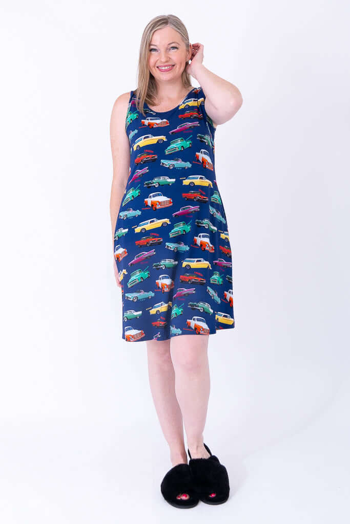 Woman in blue sleeveless dress with vibrant car print, smiling with hand on head