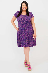 Woman modeling a purple floral A-line dress, smiling and standing front-facing