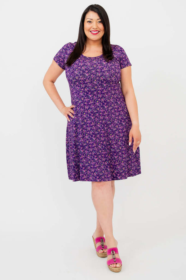 Woman modeling a purple floral A-line dress, smiling and standing front-facing