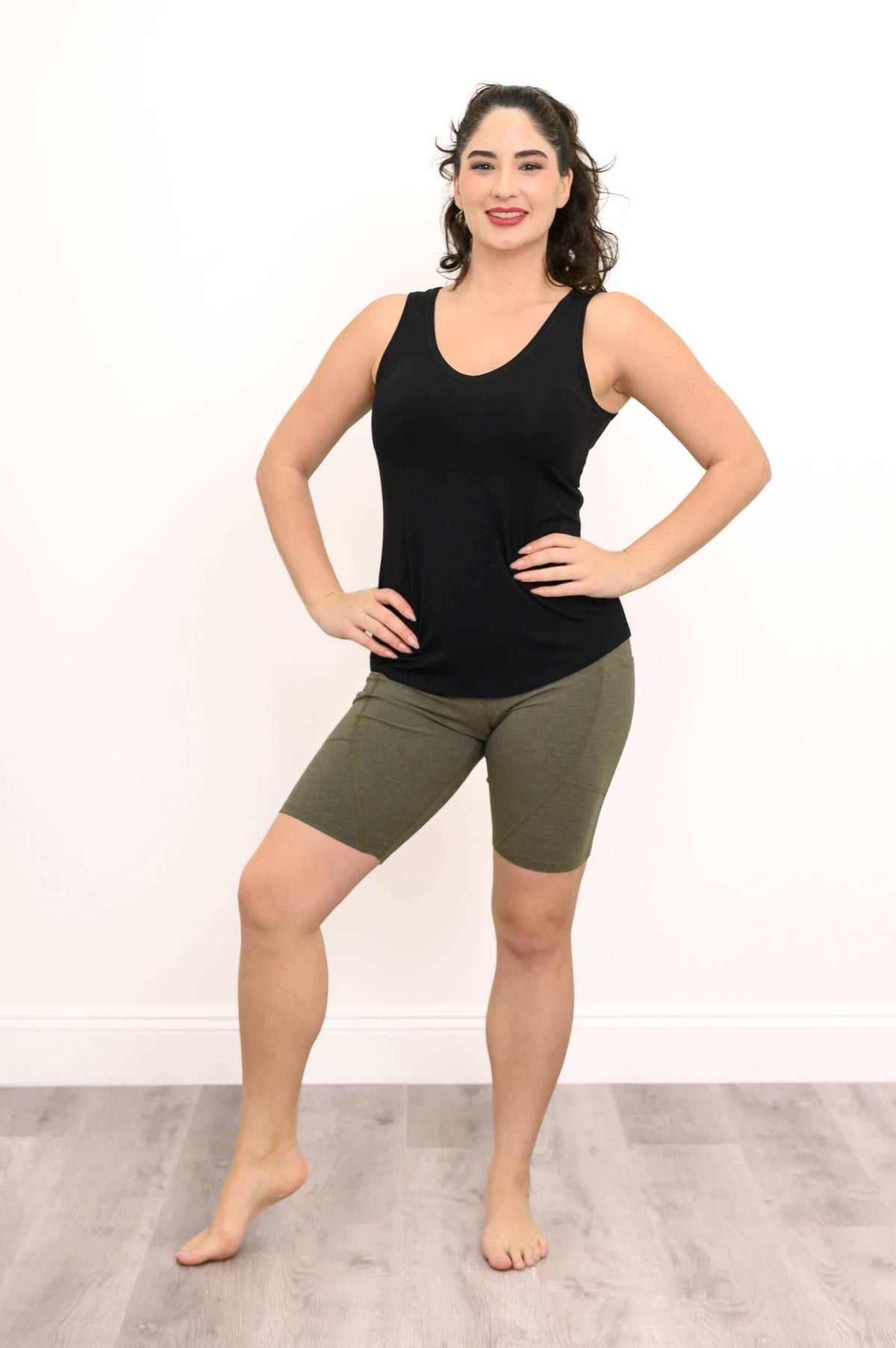 Woman in olive green bike shorts and black tank top with hands on hips, standing barefoot