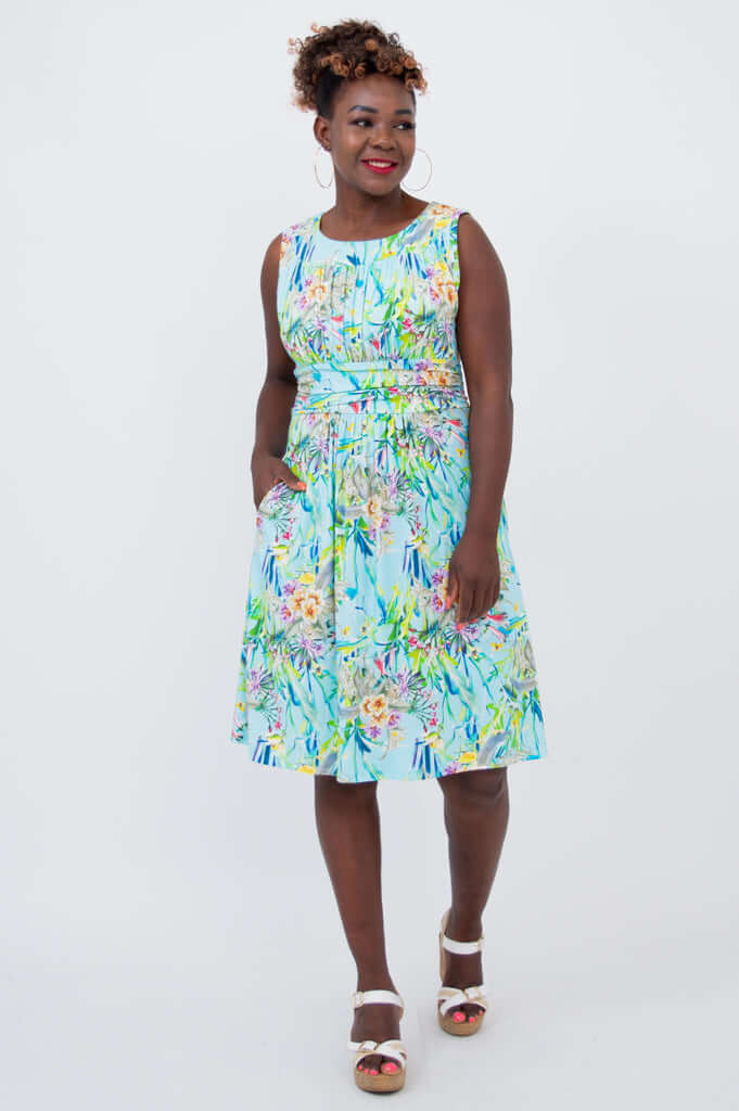 Woman smiling in sleeveless blue floral ruched dress with hands in pockets