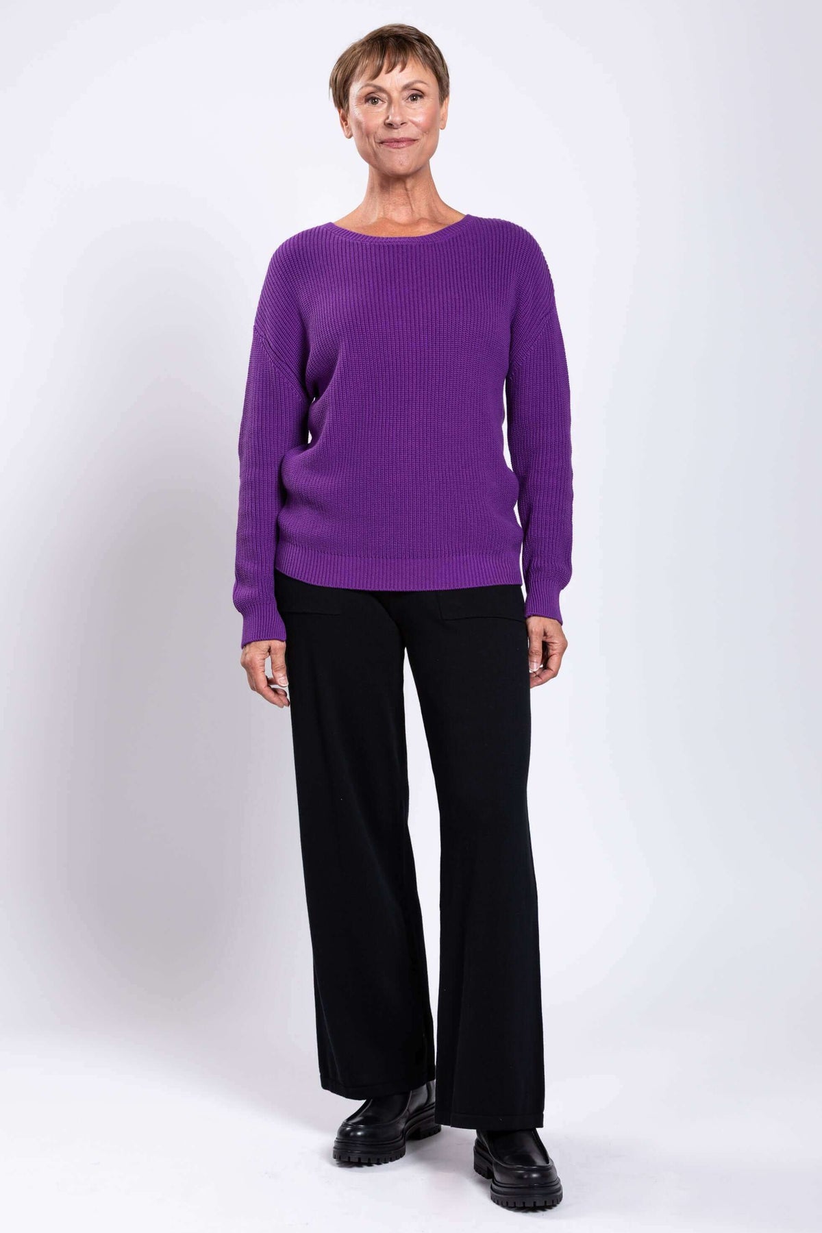 Woman standing in a purple shaker sweater and black pants, facing forward