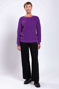 Woman standing in a purple shaker sweater and black pants, facing forward