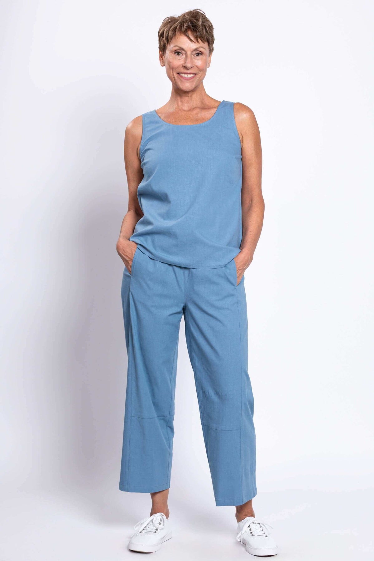 Woman in blue sleeveless top and cropped barrel-leg blue pants, hands in pockets, facing camera