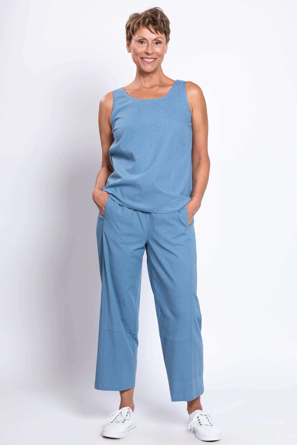 Woman in blue sleeveless top and cropped barrel-leg blue pants, hands in pockets, facing camera